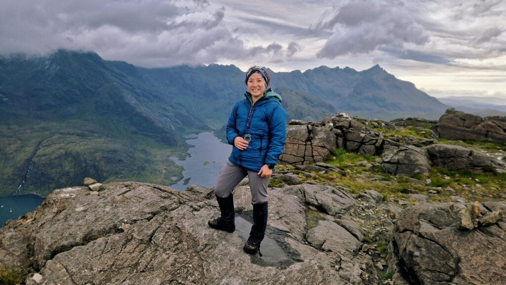 Hiker standing on a rocky mountain viewpoint with a lake and mountains behind.