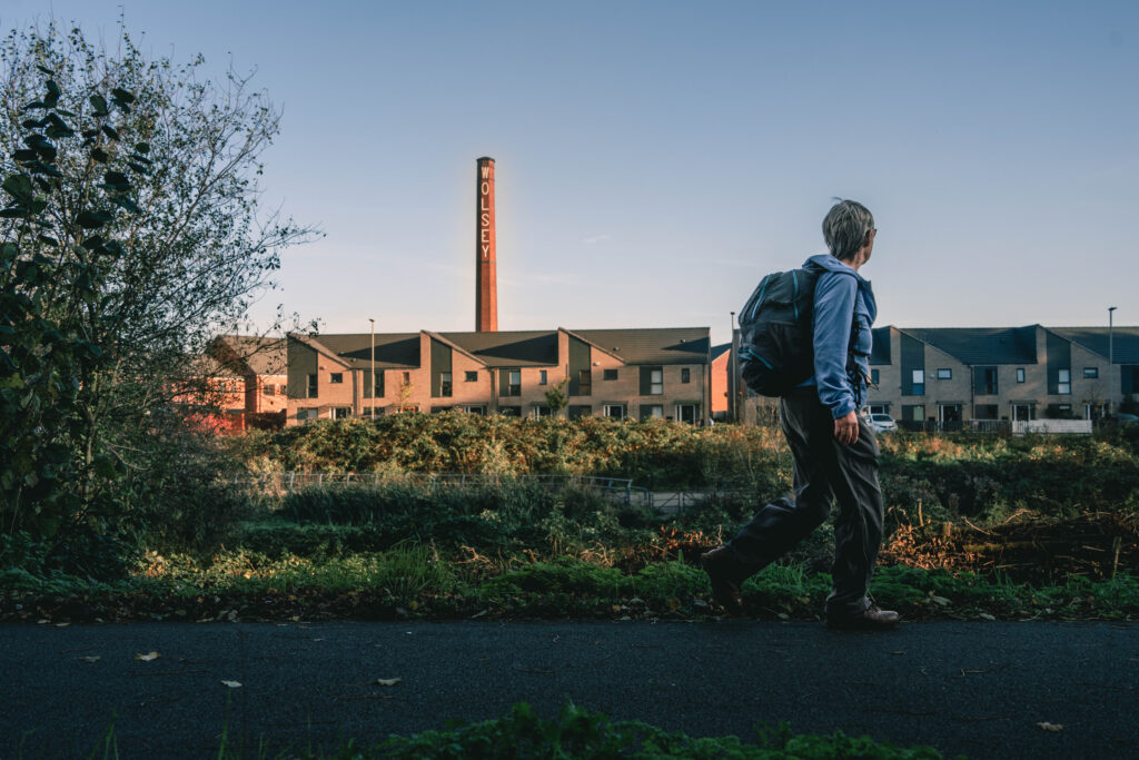 A woman with a backpack walking along a path past houses. An old and tall industrial chimney is in the backround.