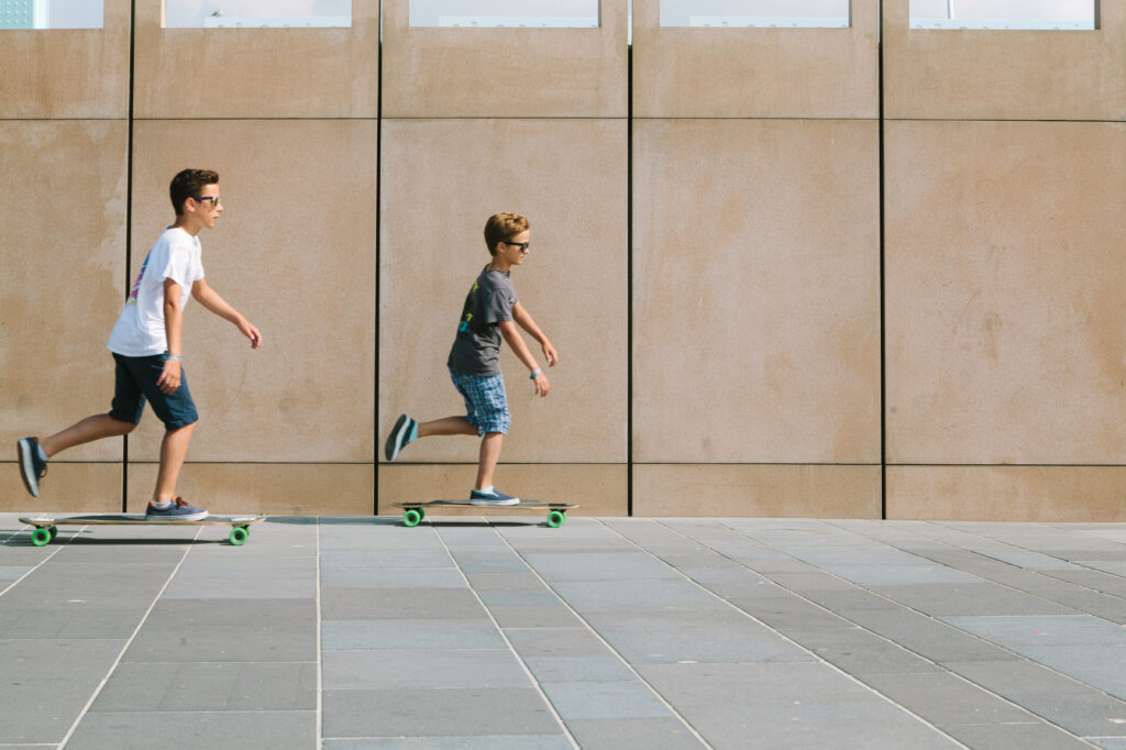 Two boys riding skateboards side by side along a paved urban walkway.