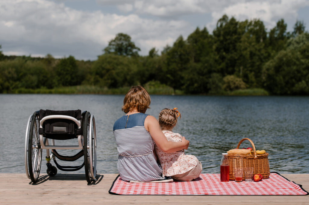 Woman and child sitting by a lake on a picnic blanket beside a wheelchair.