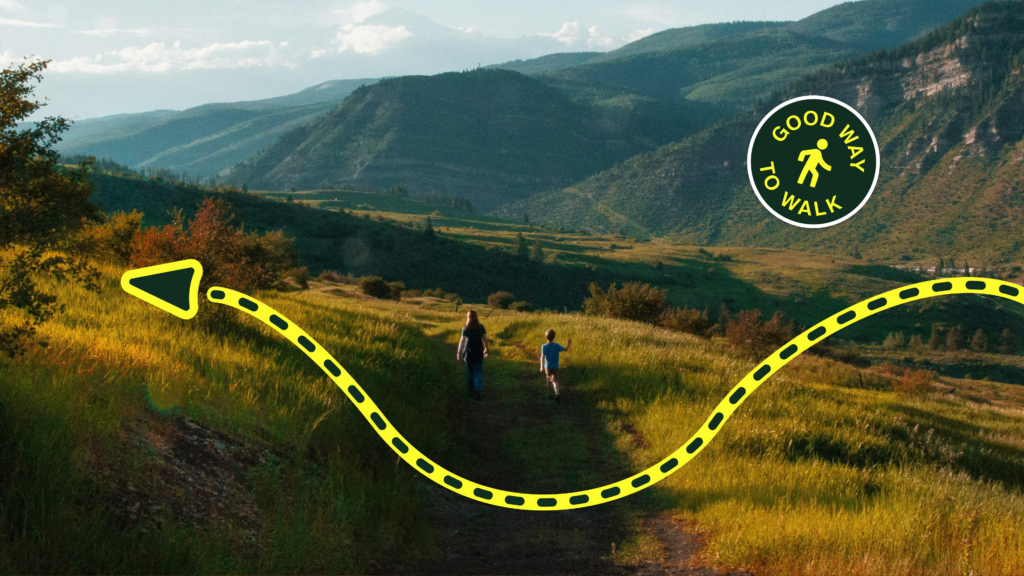 Mountain valley walking scene with two people on a trail, overlaid with a winding yellow dotted route, a navigation arrow, and a circular "Good Way to Walk" badge.