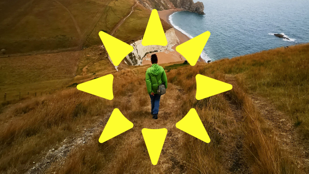 Person in a green jacket walking down a coastal path toward a beach and sea, framed by large yellow sun-ray shapes.