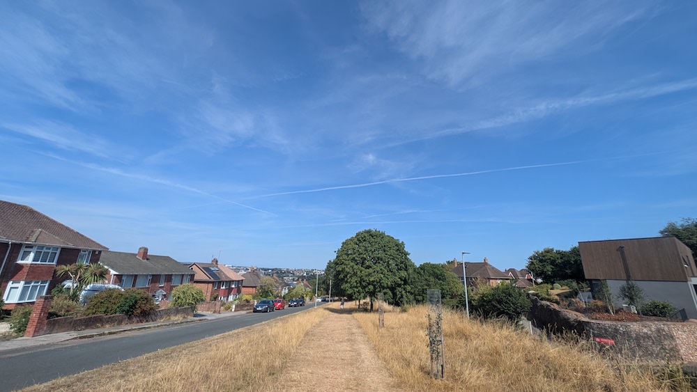 Looking from left to right we see suburban housing, a road with a few parked cars, a long straight park with a path and trees and then more housing. A person is walking on the path in the distance.