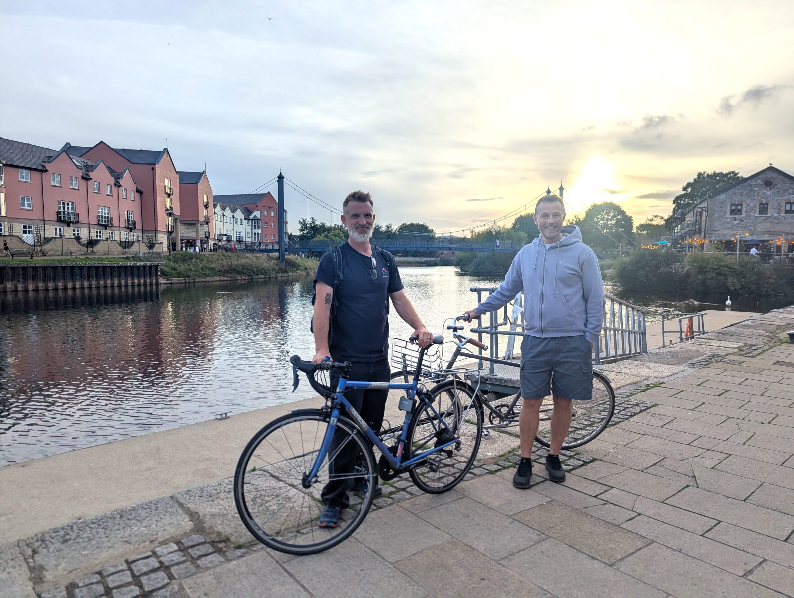Zsolt Schuller and Dan Raven-Ellison holding bikes on Exeter Quay with the River Exe in the back ground.