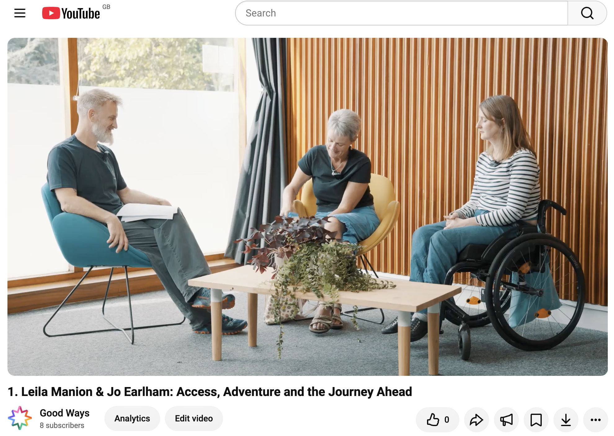 Screenshot of a YouTube video titled “Leila Manion & Jo Earlham: Access, Adventure and the Journey Ahead” on the Good Ways channel. The video shows three people seated in a bright, modern room with large windows and a wooden slatted wall. On the left, Dan Raven-Ellison, with a beard sits in a turquoise chair holding papers. In the centre, Jo Earlham, short-haired woman in a black top and blue skirt sits in a yellow chair, looking down. On the right, Leila Manion, with shoulder-length brown hair, wearing a striped top and blue jeans, sits in a black manual wheelchair. A low wooden coffee table with plants stands between them. YouTube video controls and channel information are visible below the image.